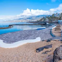 Słoneczna plaża Playa de las Américas z basenem, kolorowymi budynkami i górami w tle.