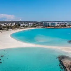 Piękna plaża Ayia Napa z turkusową wodą i białym piaskiem, otoczona hotelami i zielenią.