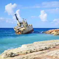Wraki statków na pafos plażach. Zardzewiały statek osiadł na mieliźnie przy skalistym wybrzeżu, woda jest turkusowa, a niebo błękitne z białymi chmurami.