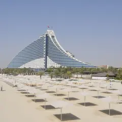 Pusta plaża z rzędami parasoli i charakterystycznym hotelem w tle. Ceny w Dubaju mogą być wysokie, ale widoki są niezapomniane.