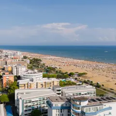 Widok z lotu ptaka na zatłoczoną bibione plażę, pełną białych parasoli i ludzi. W tle błękitne morze i nadmorskie miasteczko.