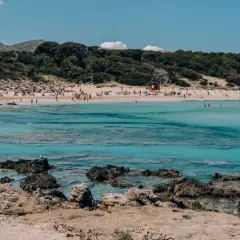 Słoneczna plaża w **Can Pastilla** z turkusową wodą, kamienistym wybrzeżem i ludźmi odpoczywającymi na piasku.