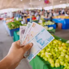 Ręka trzyma banknoty 5, 20 i 100 lir tureckich na tle targu. Widać świeże warzywa, co sugeruje ceny w Turcji.