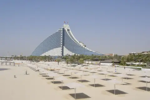 Pusta plaża z rzędami parasoli i charakterystycznym hotelem w tle. Ceny w Dubaju mogą być wysokie, ale widoki są niezapomniane.