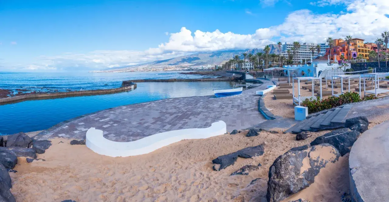 Słoneczna plaża Playa de las Américas z basenem, kolorowymi budynkami i górami w tle.