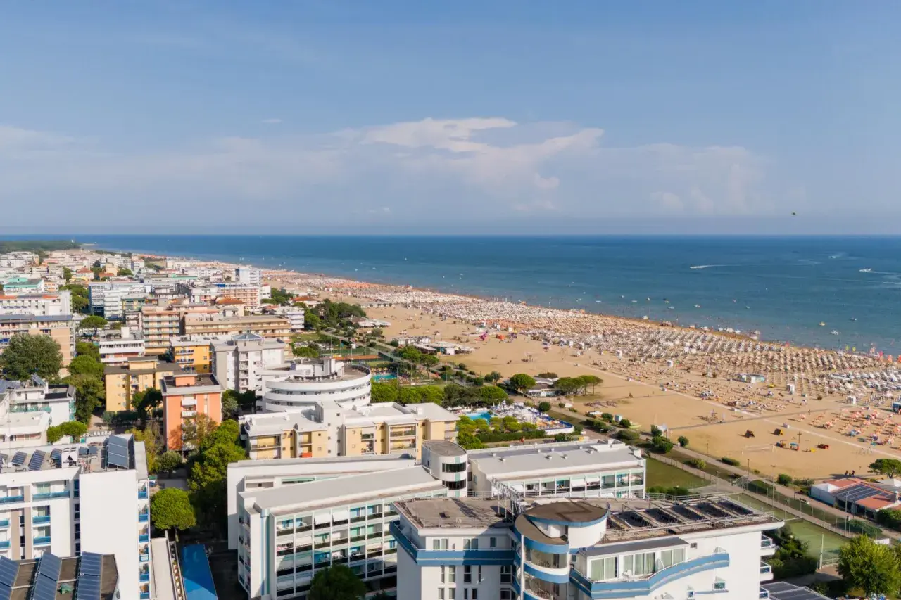 Widok z lotu ptaka na zatłoczoną bibione plażę, pełną białych parasoli i ludzi. W tle błękitne morze i nadmorskie miasteczko.