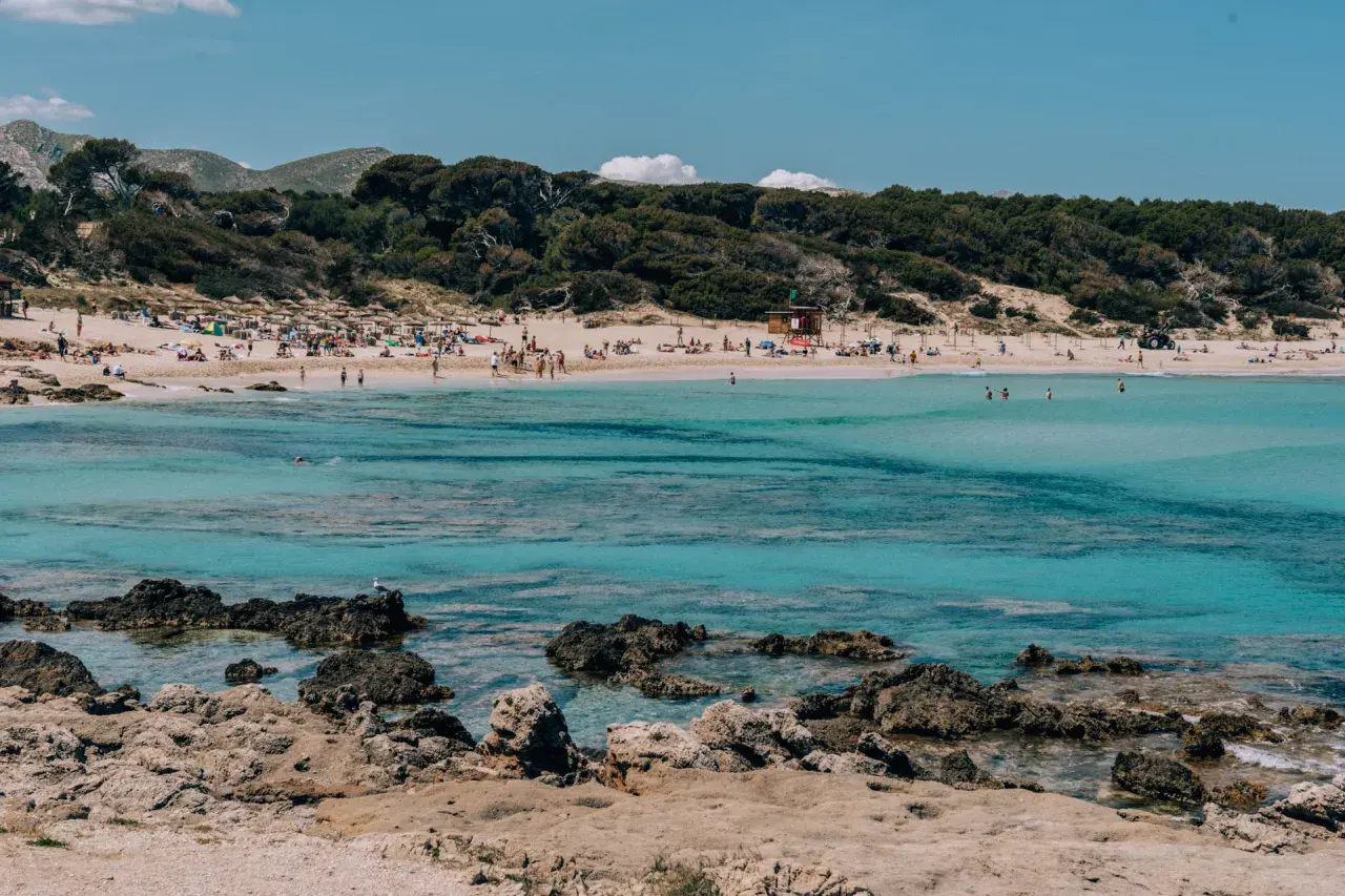 Słoneczna plaża w **Can Pastilla** z turkusową wodą, kamienistym wybrzeżem i ludźmi odpoczywającymi na piasku.