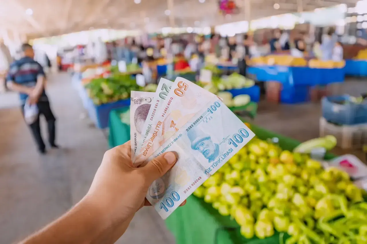Ręka trzyma banknoty 5, 20 i 100 lir tureckich na tle targu. Widać świeże warzywa, co sugeruje ceny w Turcji.