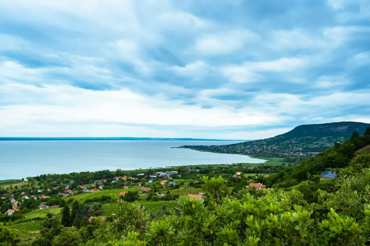 Widok na jezioro Balaton z zielonymi wzgórzami i domkami. Chmury na niebie tworzą dramatyczny krajobraz.