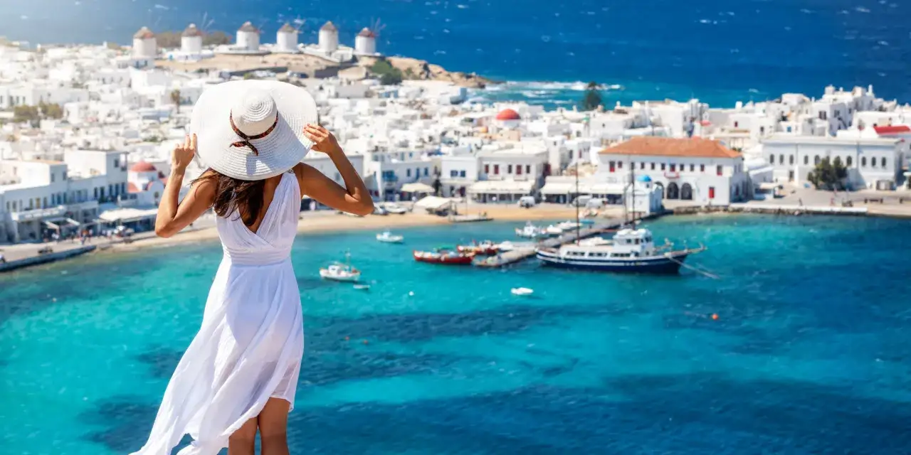 Kobieta w białej sukni podziwia widok na Mykonos. Wiatraki, białe domy i turkusowe morze. Sprawdź grecja ceny, by zaplanować wymarzone wakacje!
