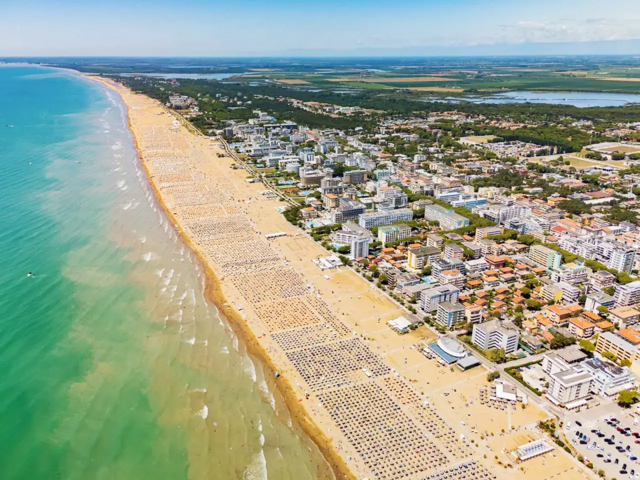 Słoneczna bibione plaża tętni życiem, pełna leżaków i turystów. W tle widać miasteczko i zieleń.