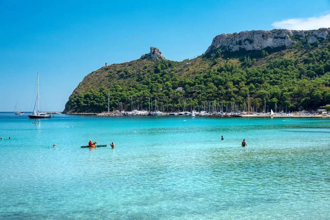 Turkusowe morze i plaża w Cagliari, stolicy Sardynii. W tle zielone wzgórza i przystań pełna jachtów.