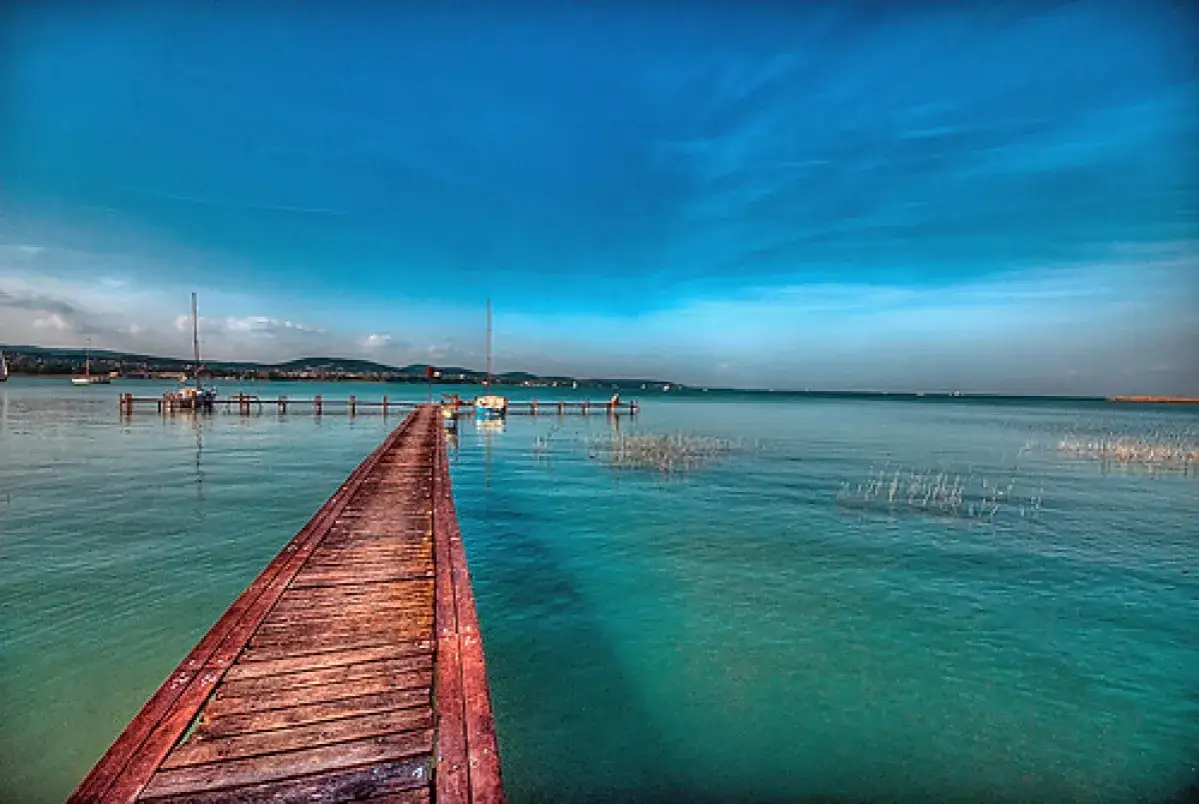 Drewniany pomost prowadzi na spokojne wody jeziora Balaton. W oddali widać żaglówki i zarys lądu.