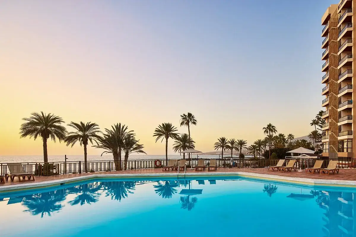 Basen z turkusową wodą i odbijającymi się palmami, hotel i ocean w tle. Idealne miejsce na wakacje na Playa de las Américas.