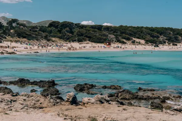 Słoneczna plaża w **Can Pastilla** z turkusową wodą, kamienistym wybrzeżem i ludźmi odpoczywającymi na piasku.